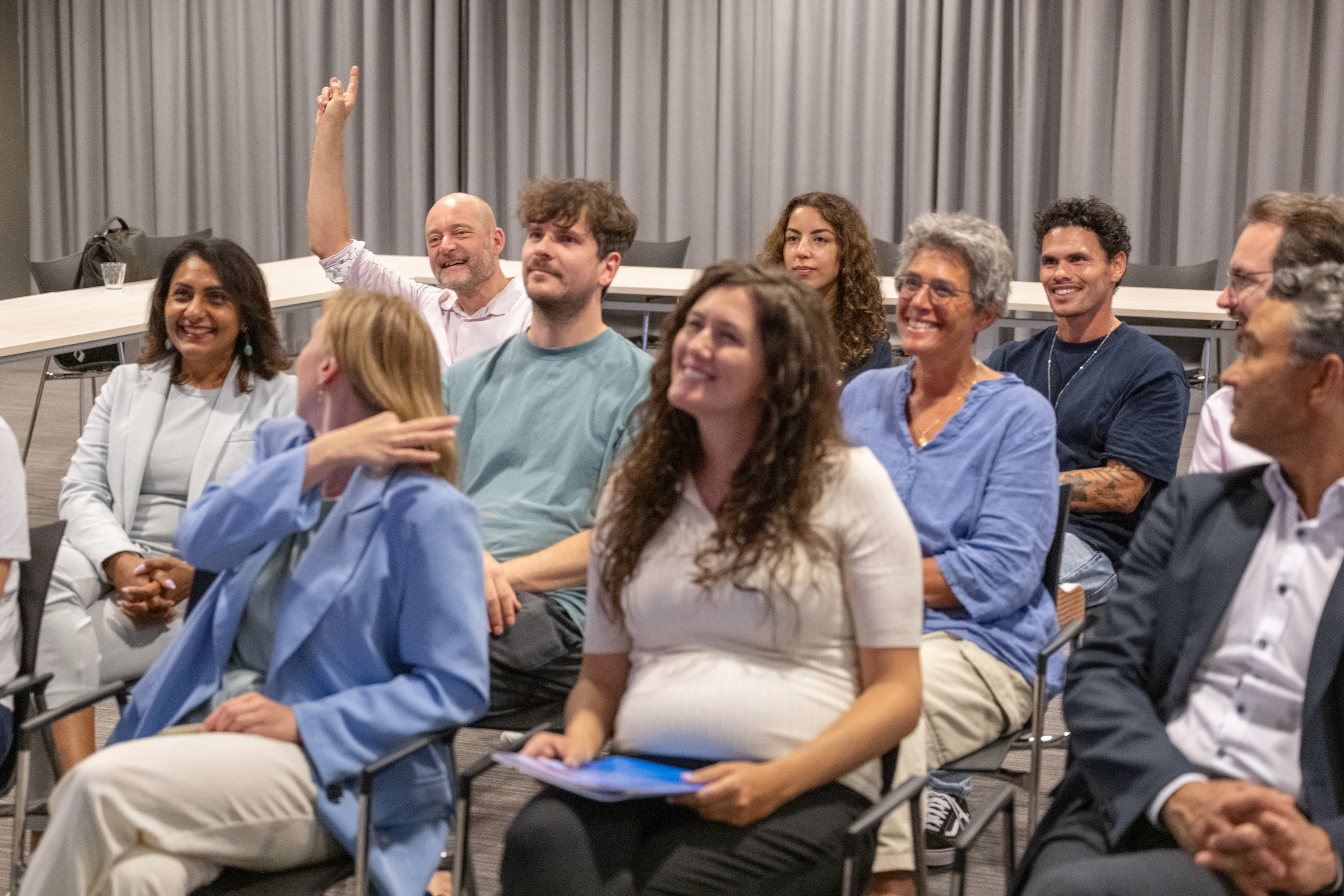Deelnemers aan een workshop. Eèn deelnemers heeft zijn vinger opgestoken omdat hij een vraag wil stellen.