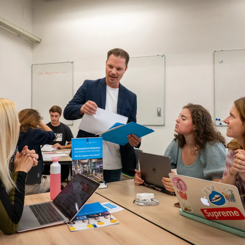 Studenten zitten in groepjes aan tafel. De spelleider deelt papieren uit
