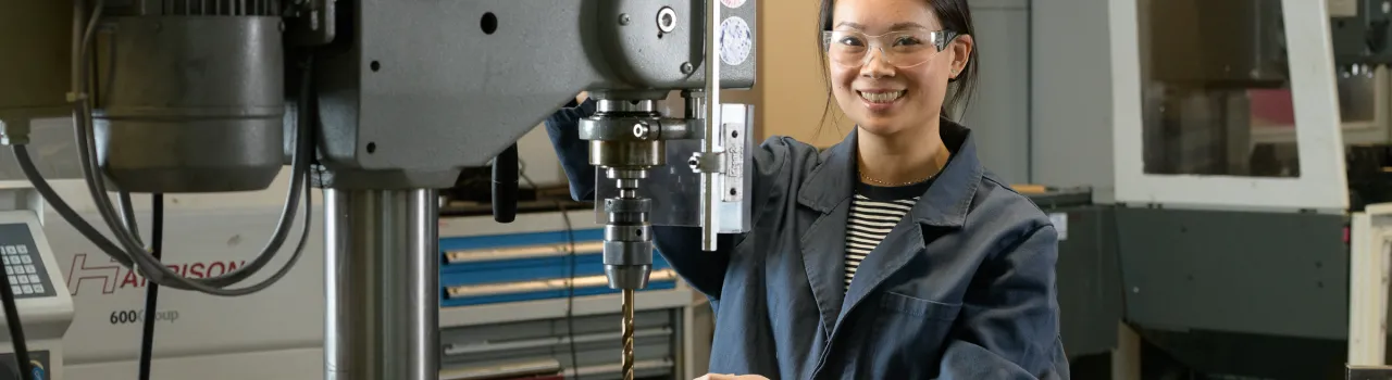 Vrouw met stofjas en beschermingsbril staat achter metaalbewerkingsmachine