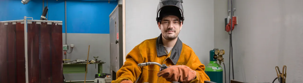 Man met lashelm op en lasapparaat in de hand staat in werkplaats